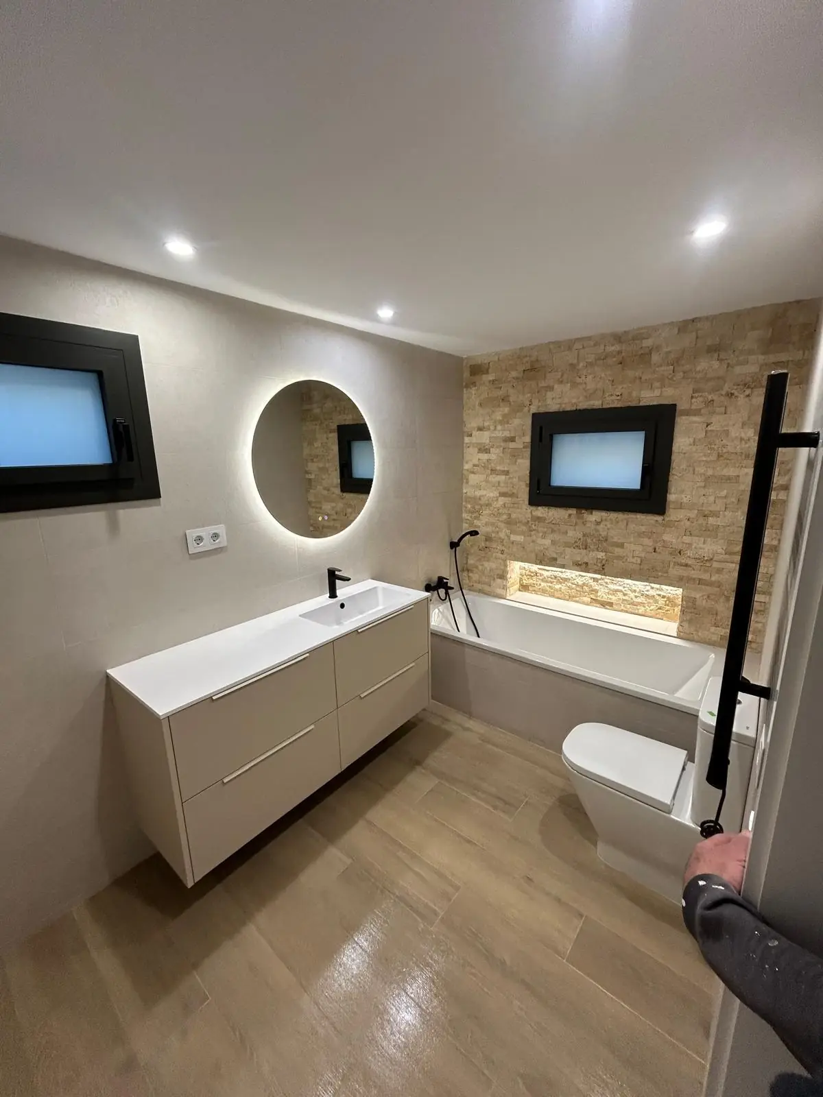 Baño reformado con espejo redondo retroiluminado, mueble suspendido y pared de piedra