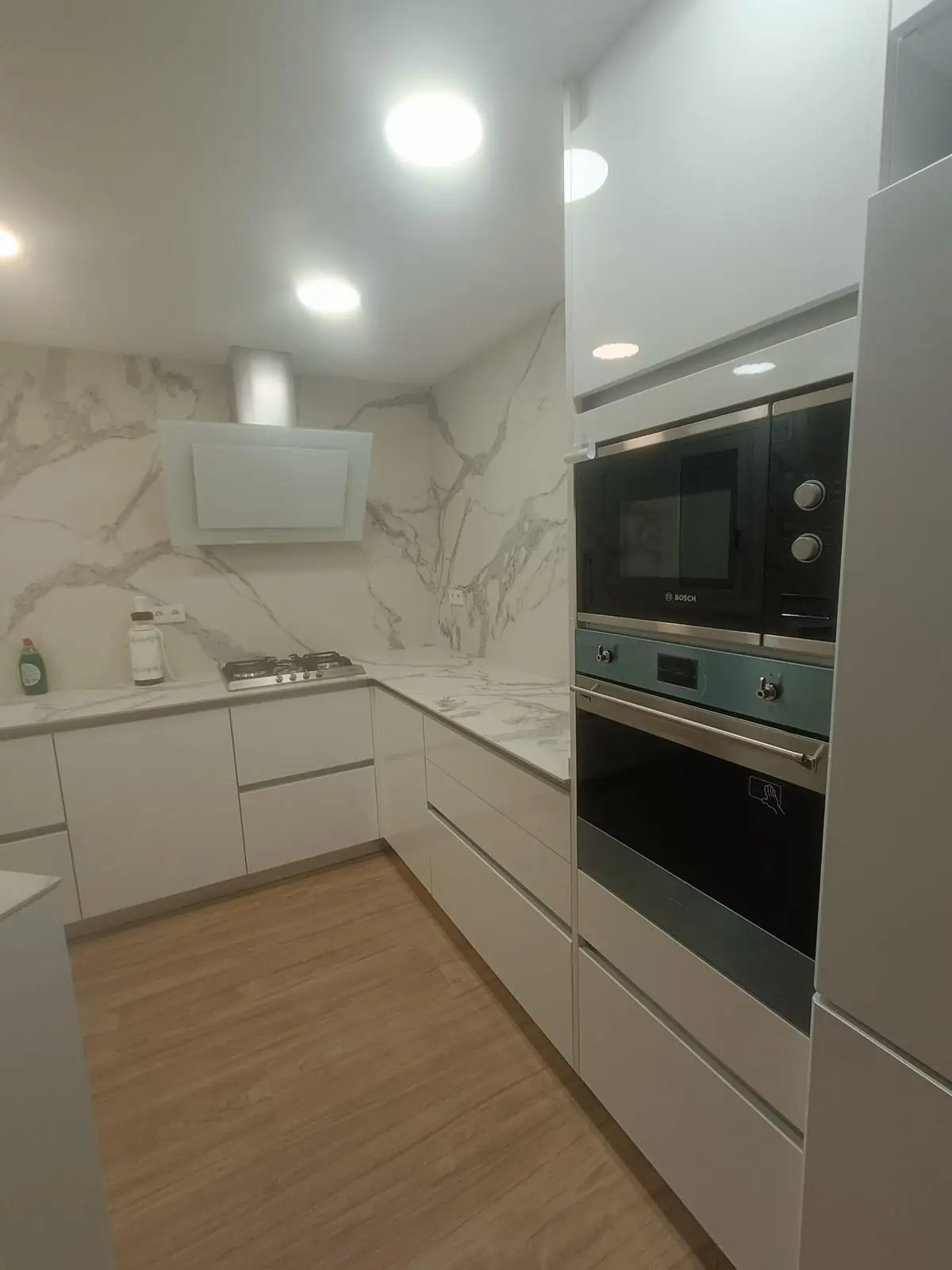 Cocina blanca en L con frontal efecto mármol y hornos en columna